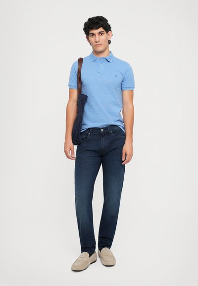Blauwe polo met korte mouwen, donkere denim jeans en beige instappers. Model houdt een marineblauwe schoudertas vast. Eenvoudig, klassiek ontwerp.