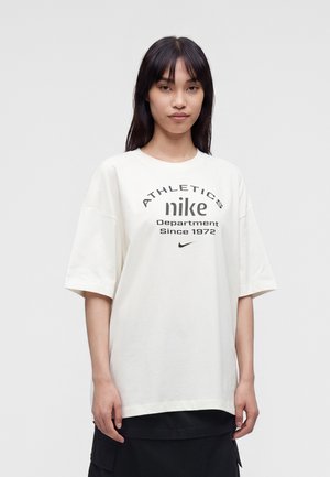 Žena s rovnými tmavými vlasmi, ktorá má na sebe biele tričko Nike s čiernym textom a malým logom Nike na hrudi, stojaca pred jednoliatym pozadím.
