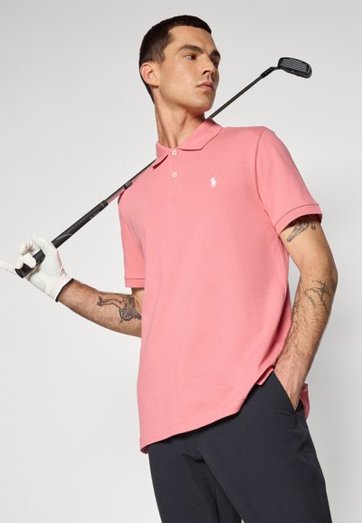 Camisa polo rosa de mangas curtas e colarinho, com um pequeno logótipo branco. O modelo segura um taco de golfe e usa uma luva branca.