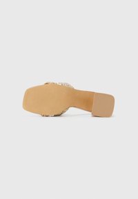 Sandale à talon avec une semelle texturée couleur tan, présentant un dessus tissé en fibres métalliques et beiges, avec un design de talon carré.