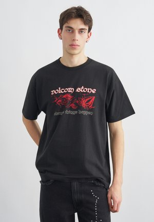 Tânăr purtând un tricou negru Volcom Stone cu grafică roșie și blugi negri cu ținte argintii, pe un fundal alb simplu.