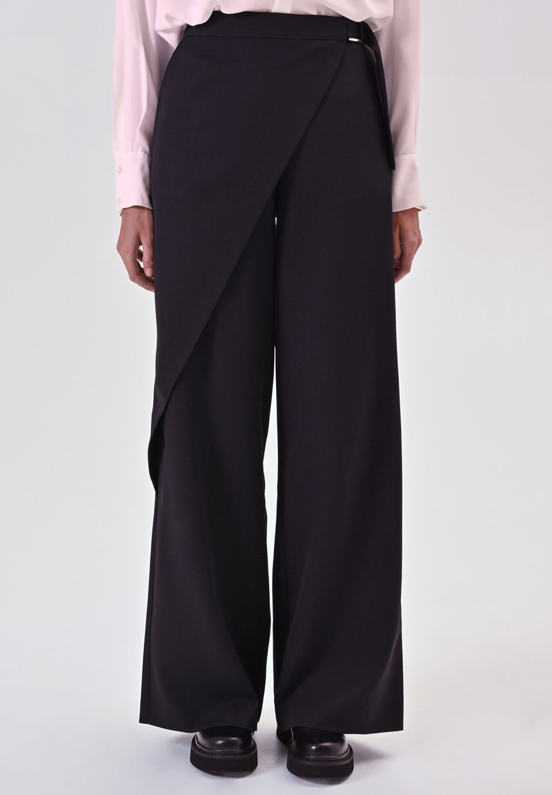 Pantaloni neri a gamba larga con un pannello frontale asimmetrico, realizzati in un tessuto liscio. Presentano un design minimalista e una finitura pulita.
