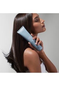 Produit de coiffage Aveda Smooth Infusion dans un tube bleu clair, tenu contre une épaule lisse. Les cheveux semblent lisses, raides et brillants.