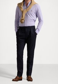 Mężczyzna w jasnofioletowej, zapiętej koszuli, z beżowym swetrem zarzuconym na ramionach, ciemnych spodniach w prążki, brązowych skórzanych mokasynach oraz zielonych skarpetkach.