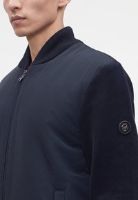Gros plan d'une personne portant une veste bomber bleu marine avec des manches en tricot et un petit logo circulaire noir sur le bras gauche.