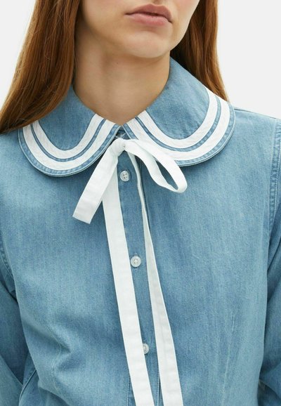 Camicia di jeans blu chiaro con un ampio colletto bianco con due righe orizzontali e un fiocco bianco al collo. Chiusura con bottoni classici.