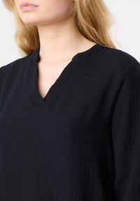Mustat, ryppyiset kankaat, joissa on V-pääntie ja hienovaraiset ompeluyksityiskohdat, joissa lukee "GEASTIE". Pehmeä pinta ja rento istuvuus.