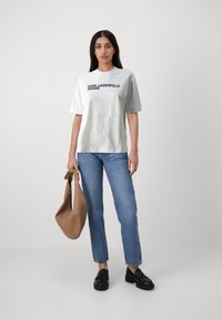 Camiseta blanca de algodón con la impresión en negro "KARL LAGERFELD JEANS", combinada con jeans rectos de color azul claro y mocasines negros. Lleva un bolso de hombro marrón.