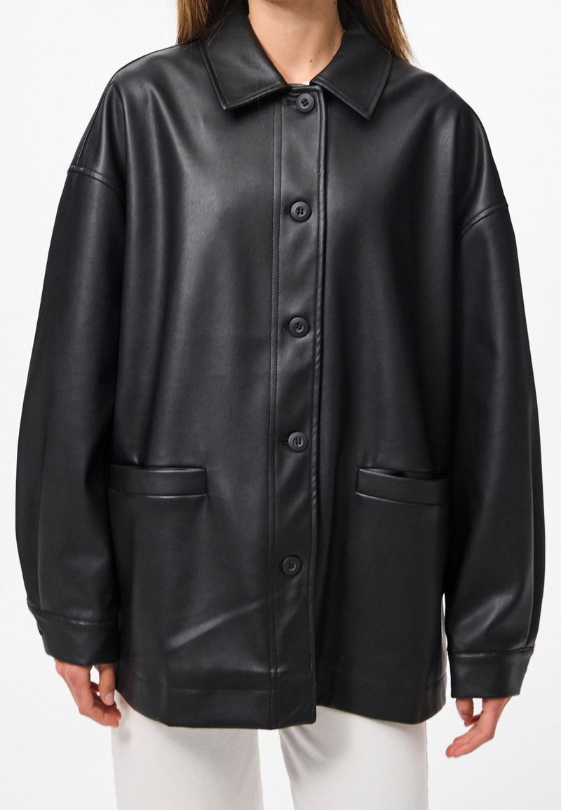 Femme portant une veste en cuir noire ample boutonnée, avec col et poches avant, associée à un pantalon blanc.
