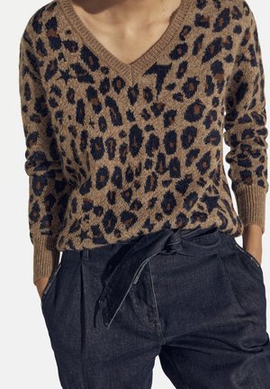 Pull à col en V imprimé léopard marron associé à un pantalon en denim foncé taille haute avec ceinture nouée et mains dans les poches.
