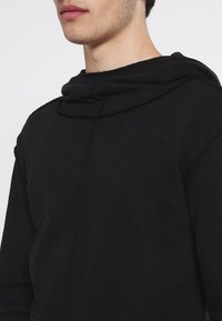Sudadera con capucha negra hecha de tejido texturizado, con un escote drapeado y un diseño minimalista. Costuras visibles en las costuras.