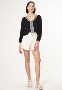 Schwarze Bluse aus strukturiertem Stoff mit einem weißen gemusterten Ausschnitt, kombiniert mit weißen hochgeschnittenen Shorts und beigen Plateausandalen.