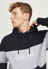 Hoodie i sort og grå, med et logo foran. Materialet har en glat tekstur med kontrasterende farveblokke og snor.