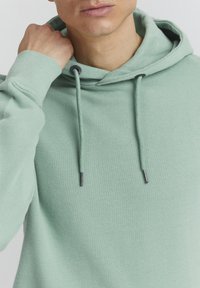 Mintgroene hoodie met een gestructureerd oppervlak, koordjes met zwarte uiteinden en een ontspannen pasvorm. Kap en geribbelde manchetten zijn ook zichtbaar.