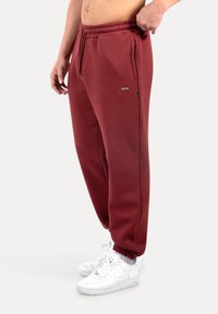 Burgundské tepláky s elastickým páskem, bočními kapsami a zúženými manžetami, doplněné bílými teniskami. Hladká textilie.