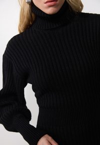 Pull à col roulé noir en maille côtelée, présentant un design ajusté, des manches longues et un tissu texturé. Les détails incluent un côtelé prononcé et un col haut.