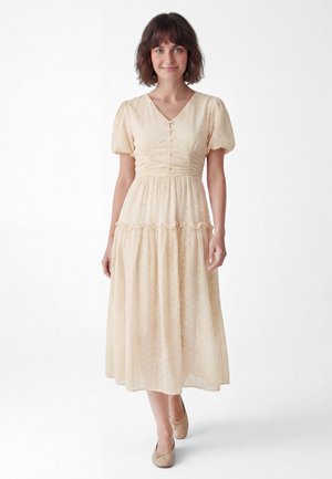 Frau mit kurzen braunen Haaren, die ein beiges, strukturiertes Midikleid mit Puffärmeln und beige Ballerinas trägt, steht vor weißem Hintergrund.
