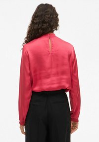 Blusa a maniche lunghe in raso fucsia brillante, con schiena a chiave e polsini arricciati. Abbinata a pantaloni neri, che presentano una texture liscia.