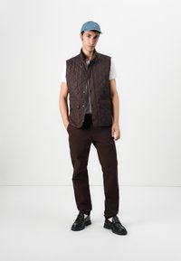 Καφέ ραμμένος γιλέκο με ψηλό γιακά, μπροστινά κουμπιά και δύο πλάγιες τσέπες, φορεμένο πάνω από λευκό t-shirt και συνδυασμένο με καφέ παντελόνι και μαύρα παπούτσια.