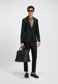 Zwarte op maat gemaakte suit met een bruine shirt, draagt een zwarte leren tas. Glad van textuur, slank ontwerp, aangesloten vorm, glanzende schoenen.