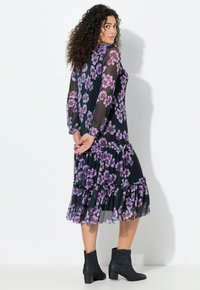 Frau mit lockigem Haar, die ein schwarzes, durchscheinendes Kleid mit lila Blumenmuster und schwarze Knöchelstiefel trägt, steht mit hinter dem Rücken verschränkten Händen.