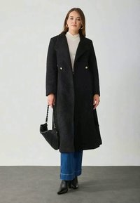 Dlhý čierny kabát s textúrovanou látkou, dvojradové zlaté gombíky a bočné vrecká. Nosený cez krémový top a kombinovaný s modrými džínsami.