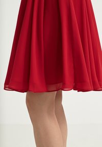 Jupe évasée rouge longueur genou avec un tissu souple drapé sur les jambes nues d'une personne sur fond blanc uni.