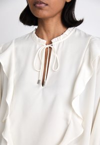 Balta blūze ar viļņotu dizainu, apaļu kakla izgriezumu un saites aizdari. Raksturīga gluda tekstūra un smalka spīduma.