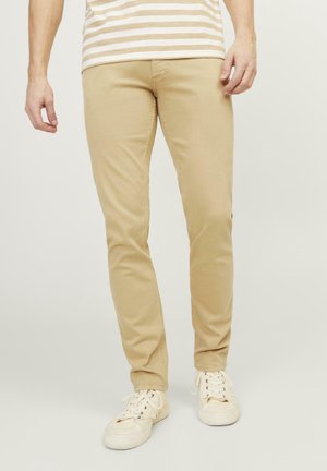 Homme portant un pantalon beige slim et des baskets blanches, debout devant un fond clair uni.