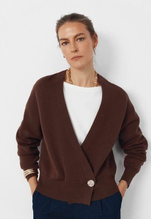 Brun strikket cardigan med omslagsdesign, med en fremtrædende sølvknap, ribstrikkede ærmekanter og en afslappet pasform, båret over en hvid top.