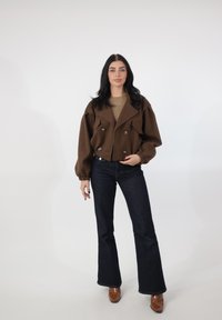 Brązowa krótka marynarka dwurzędowa z dużymi guzikami, bufiaste rękawy, w połączeniu z ciemnymi rozszerzanymi dżinsami i brązowymi butami na obcasie.