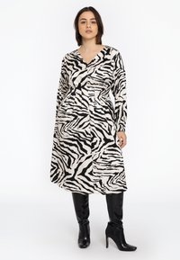 Zebra-Print-Kleid in Schwarz und Creme mit Wickeldesign, langen Ärmeln und taillierter Taille, kombiniert mit kniehohen schwarzen Stiefeln.