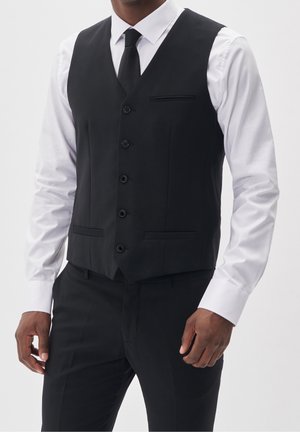 Mężczyzna ubrany w czarną, dopasowaną kamizelkę, czarny krawat, czarne spodnie i białą koszulę, stojący na jednolitym tle.