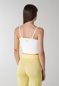 Top blanco corto con tirantes finos, textura acanalada ajustada y detalles de hardware en tono dorado ajustables. Combinado con pantalones ajustados de color amarillo claro.
