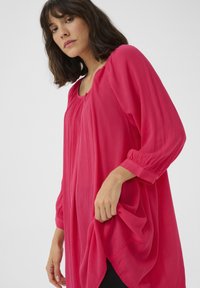 Haut fluide à manches longues de couleur rose vif, présentant un tissu texturé, un col arrondi et des détails froncés aux épaules.
