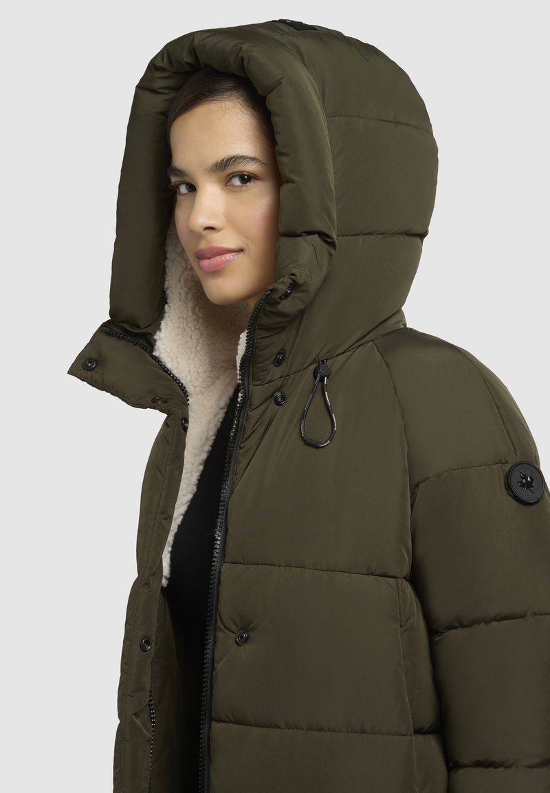 Olivgrüne Pufferjacke mit großer Kapuze, Fleece-Futter, sichtbarem Reißverschluss und Druckknöpfen. Hat ein Seitenlogo-Patch und einen Kordelzug.