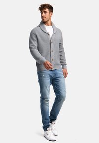 Grauer gestrickter Cardigan mit einem Schalkragen, ausgestattet mit großen Holzknöpfen, kombiniert mit ausgefransten hellblauen Jeans und weißen Turnschuhen.
