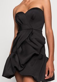 Zwarte strapless jurk met een aansluitend lijfje en geplooid ontwerp. De stof is glad met een rijke textuur, voorzien van een uitlopende zoom en draperieaccenten.