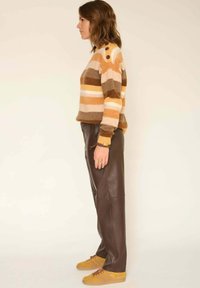 Pull rayé dans des tons chauds de marron, orange et crème, avec des accents de boutons sur l'épaule, associé à un pantalon en simili cuir marron et des chaussures jaunes.