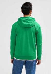 Grøn hættetrøje lavet af blødt stof med kængurulomme, ribstrikkede manchetter og kant. Kombineret med blå jeans.