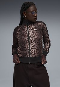 Γυναίκα με πλεξούδες και γυαλιά φοράει μια ζακέτα Puma με φερμουάρ και λεοπάρ μοτίβο και σκουρόχρωμο παντελόνι, στέκεται μπροστά σε γκρίζο φόντο.