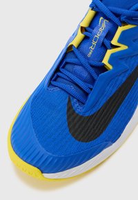 Baskets de sport bleues avec une tige en mesh texturé, des accents jaunes et un logo Nike noir. La semelle est blanche avec un motif strié.