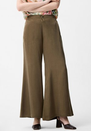 Vrouw draagt olijfgroene broek met hoge taille en wijde pijpen met een trekkoord in de taille en bruine schoenen met blokhak, staand tegen een witte achtergrond.