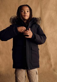 Čierna parka s kožušinovým lemom na kapucni, prednými gombíkmi a dvoma bočnými vreckami. Model má na sebe béžové cargo nohavice. Pozadie je svetlohnedé.