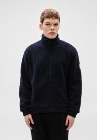 Pehme sinise värvusega fliisist lukuga jakk, millel on kõrge krae, tekstuuriga pind ja soonikuga varrukasäärised. Ees kulgeb tõmbeluku sulgur.