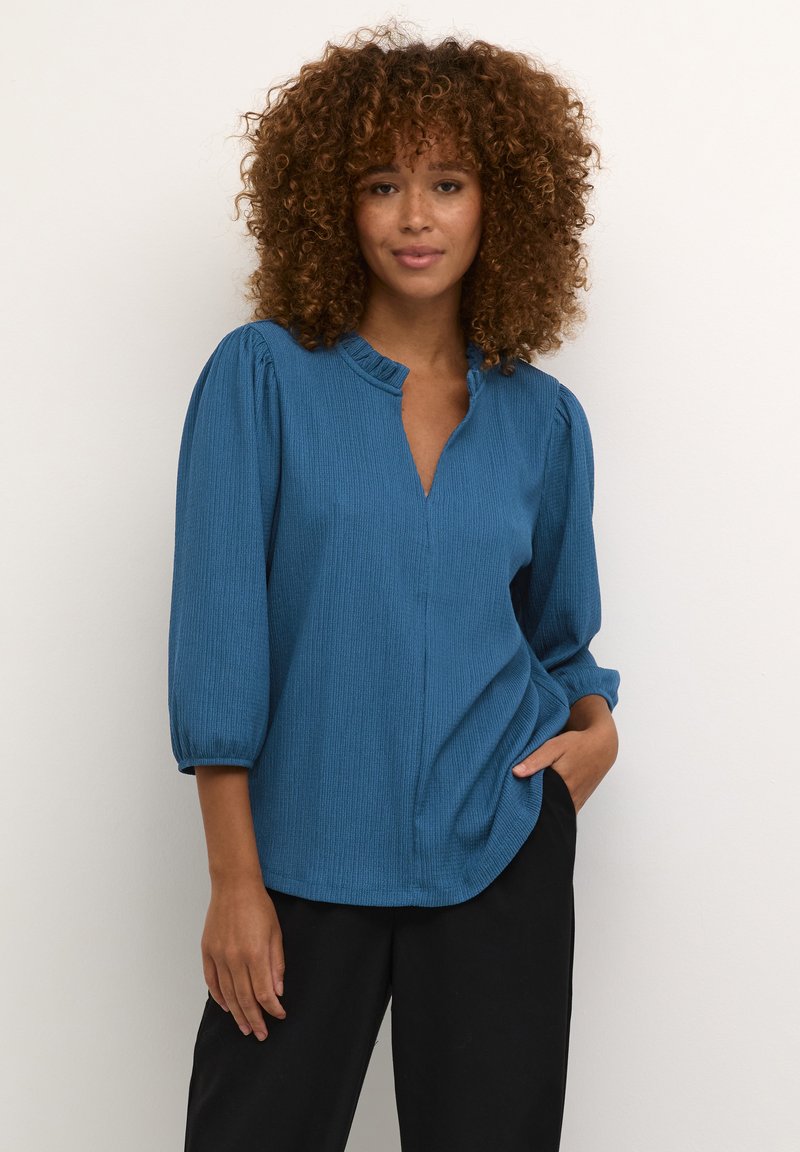 Blouse bleue côtelée avec un col à volants, des manches trois-quarts et une coupe décontractée, rentrée dans un pantalon noir. Texture lisse et éclat subtil.