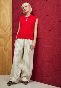 Red sleeveless knit top, beige pinstriped wide-leg pants with logo, and animal-print shoes against a hexagonal textured background.