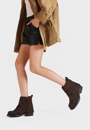 Stivaletti in suede marrone, con zip laterale, punta tonda e suola texturizzata, abbinati a pantaloni corti neri e una giacca marrone chiaro.