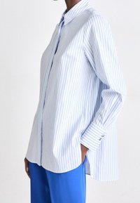 Osoba nosící volnou košili s dlouhými rukávy, světle modro-bílé pruhované, která není zastrčená, přes jasně modré kalhoty, ruka v kapse.