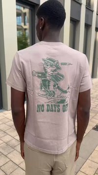 Camiseta de algodón color beige con un gráfico verde de un gato con gafas de sol y un sombrero, con el texto "NO HAY DÍAS LIBRES" debajo.